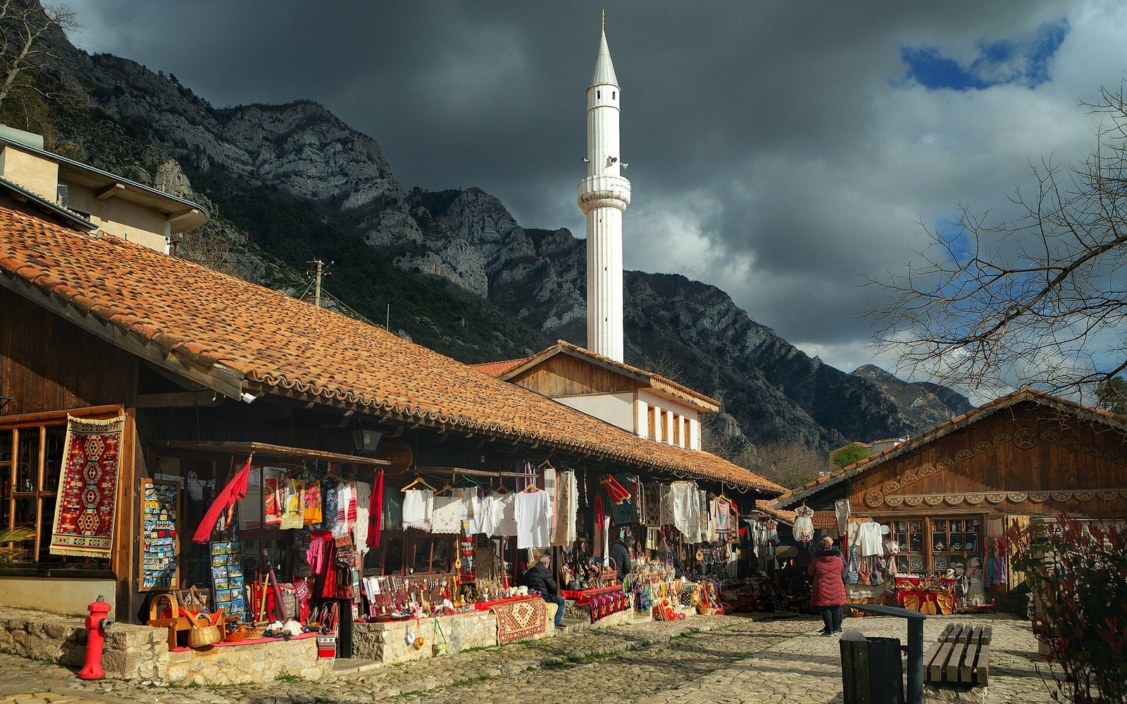 A covered bazaar street of timber shopfronts below a white minaret and a limestone ridge