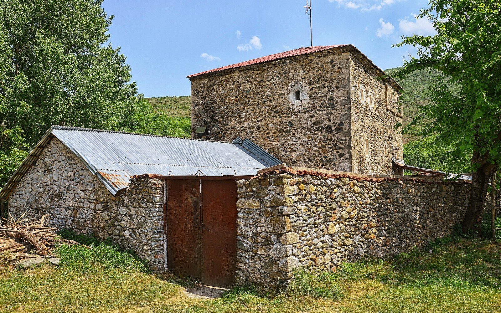 A stone tower-house behind a rubble wall and a low outbuilding, wooded hills behind